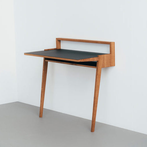 The Bachmann Desk by Das kleine b is a minimalist wooden desk with two legs and a sleek black surface, mounted on a white wall. It includes a narrow shelf above the main workspace, offering a modern and compact design perfect for contemporary office spaces.