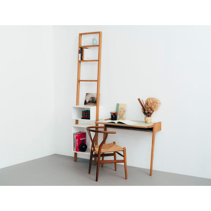 A minimalist workspace showcases a Bachmann Desk by Das kleine b and a chair. Books, a notebook, and stationery sit on the solid oak desk. A ladder-style shelf leans against the wall with books and decor, exuding simplicity and organization.