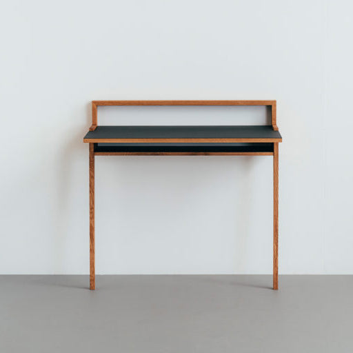 The Bachmann Desk by Das kleine b is a minimalist solid oak piece featuring clean lines and a dark grey linoleum top. It stands against a light grey wall and floor, with a slight elevation at the back, offering a sleek modern look.