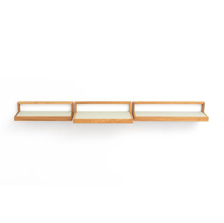 The Buck by Das kleine b is a minimalist wooden wall shelf with three connected rectangular sections. It features flat surfaces and back panels for continuous storage, has a light natural finish, and is elegantly mounted on a white wall to meet adaptable furniture needs.