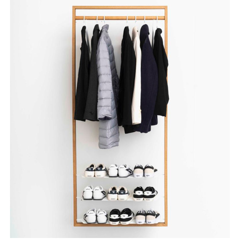 The Gardero-b by Das kleine b is a minimalist clothing rack that resembles a sleek hall stand against a white wall, showcasing several black, gray, and white jackets and coats on hangers. Below, three shelves function as shoe racks with neatly arranged pairs of black and white shoes.
