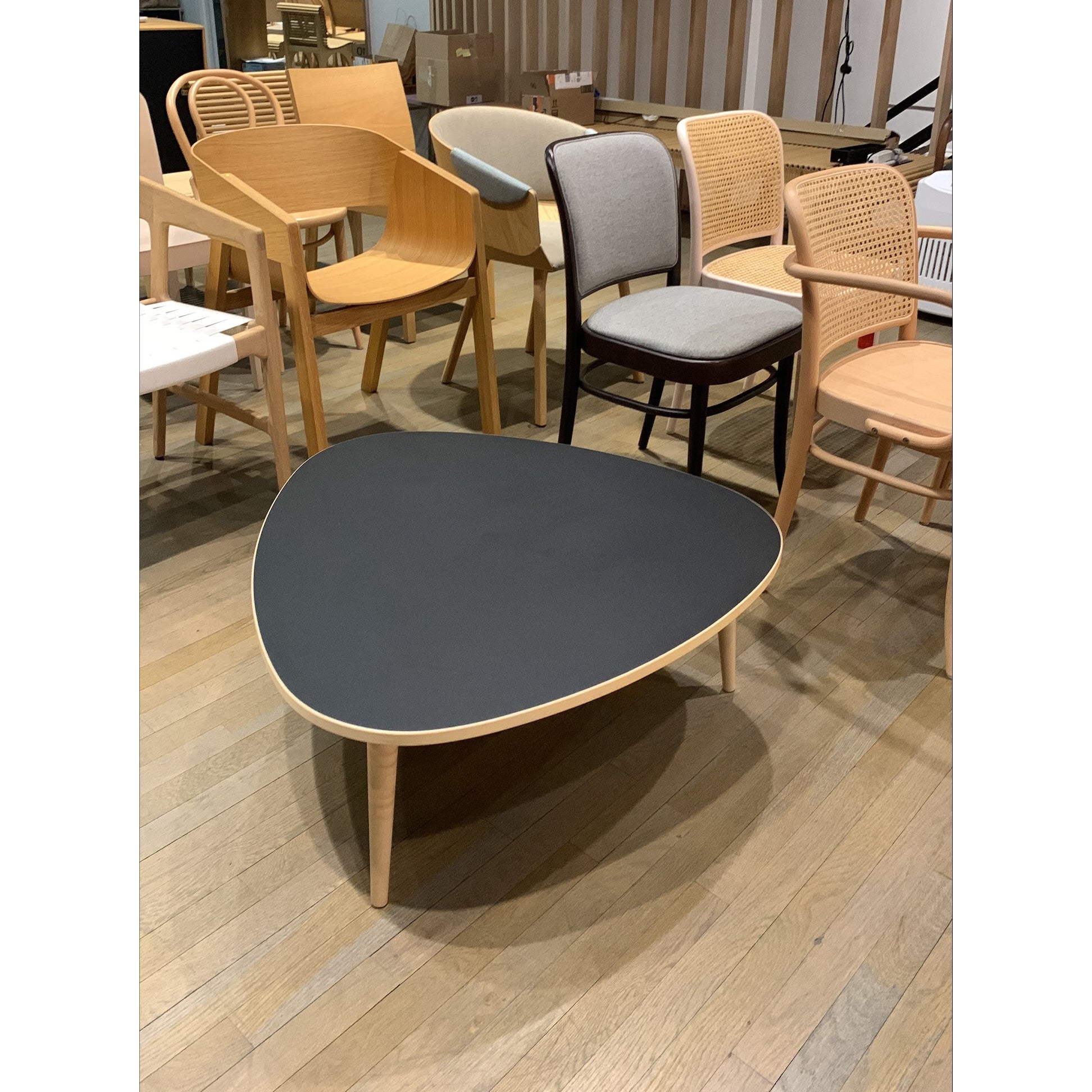 A furniture showroom showcases various chairs with wooden and woven designs. The standout piece is the Drei-Rund Coffee Table by WB Form, featuring a triangular black tabletop with wooden legs that echo kidney-shaped curves. The wooden floor highlights more exquisite pieces in the background.