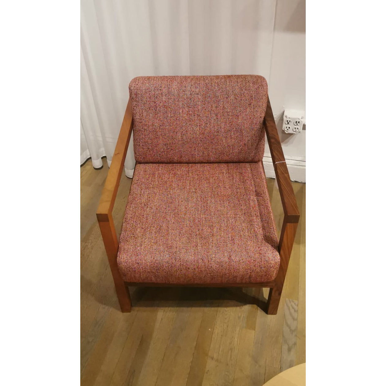 The EC01 BYRON - Sale by E15 is an elegant modern smoking chair with a wooden frame and red, speckled upholstery on a wooden floor. Its framed by white curtains, with discreet electrical outlets in the background.