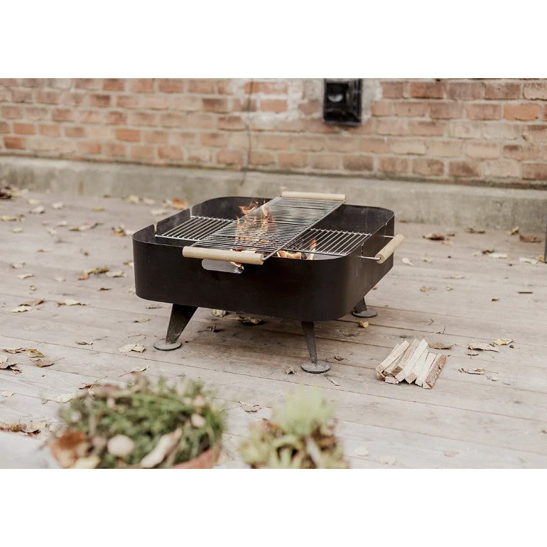 The Back to Fire Fire Pit by Egoe, a black rectangular grill with a fire-resistant grate, rests on the wooden deck. Flames flicker among scattered leaves, with firewood stacked nearby and a brick wall evoking traditional fireplace charm in the backdrop.