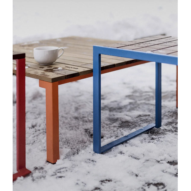 An Egoe Bistrot Table with colorful metal legs and a white cup sits on snowy ground. The table, made from tropical wood with a blue and orange frame, contrasts beautifully against the pristine snow around it.