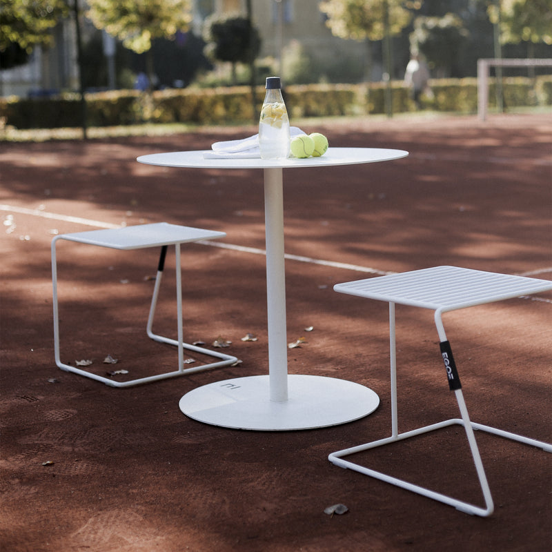 The Spulka Table by Egoe, with its durable all-metal body, holds a water bottle, folded towel, and two tennis balls on the clay tennis court. Two white metal chairs complement this minimalist round white table, surrounded by flourishing trees and greenery for a serene scene.