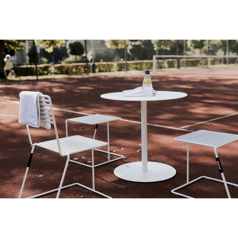 On a red tennis court, the Spulka Table by Egoe, accompanied by a bottle and towel, is encircled by two durable white all-metal chairs. The trees and net create a serene backdrop.