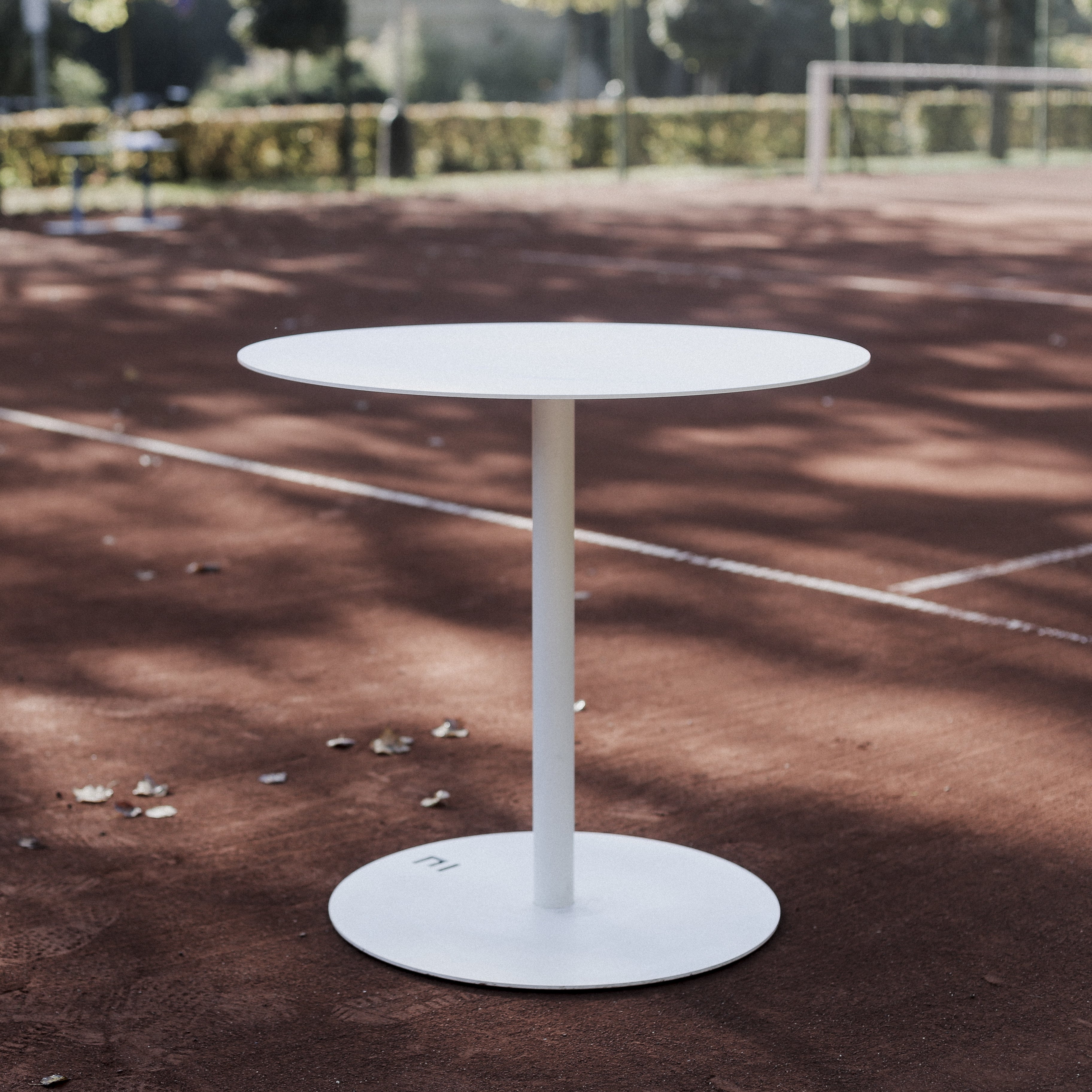 The Spulka Table by Egoe, featuring a small, round white all-metal design with a single central support, stands elegantly on a red clay tennis court as sunlight filters through nearby trees, casting soft shadows and creating an eye-catching contrast against the vibrant surface.