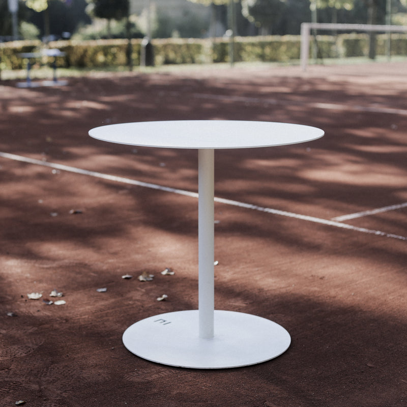 The Spulka Table by Egoe, featuring a small, round white all-metal design with a single central support, stands elegantly on a red clay tennis court as sunlight filters through nearby trees, casting soft shadows and creating an eye-catching contrast against the vibrant surface.