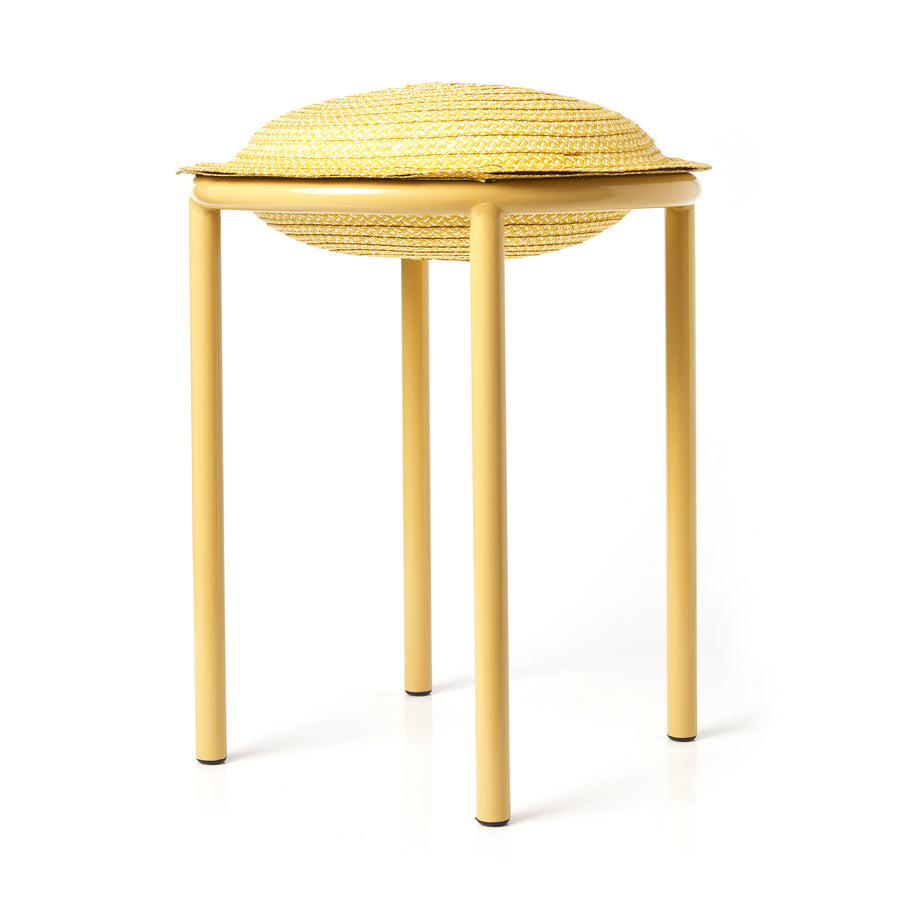 The Ames Cana Stool is a unique piece with vivid colors, featuring three yellow legs and a round woven seat that resembles a straw hat with a brim, casting a subtle shadow on the white surface below.