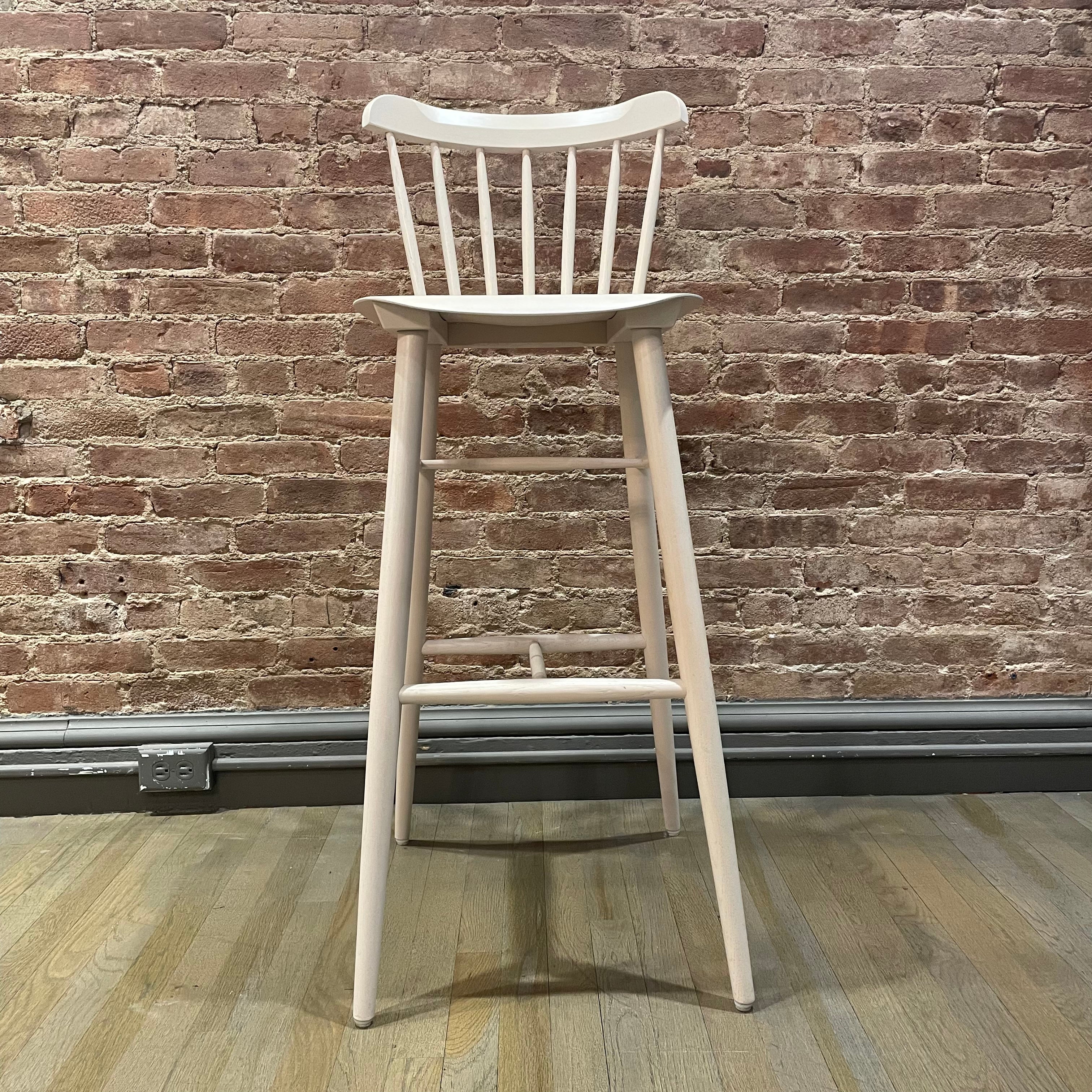 The Barstool Ironica - Sale by TON, featuring a tall oak design with a rounded backrest and spindles, stands on a wooden floor against a rustic brick wall.