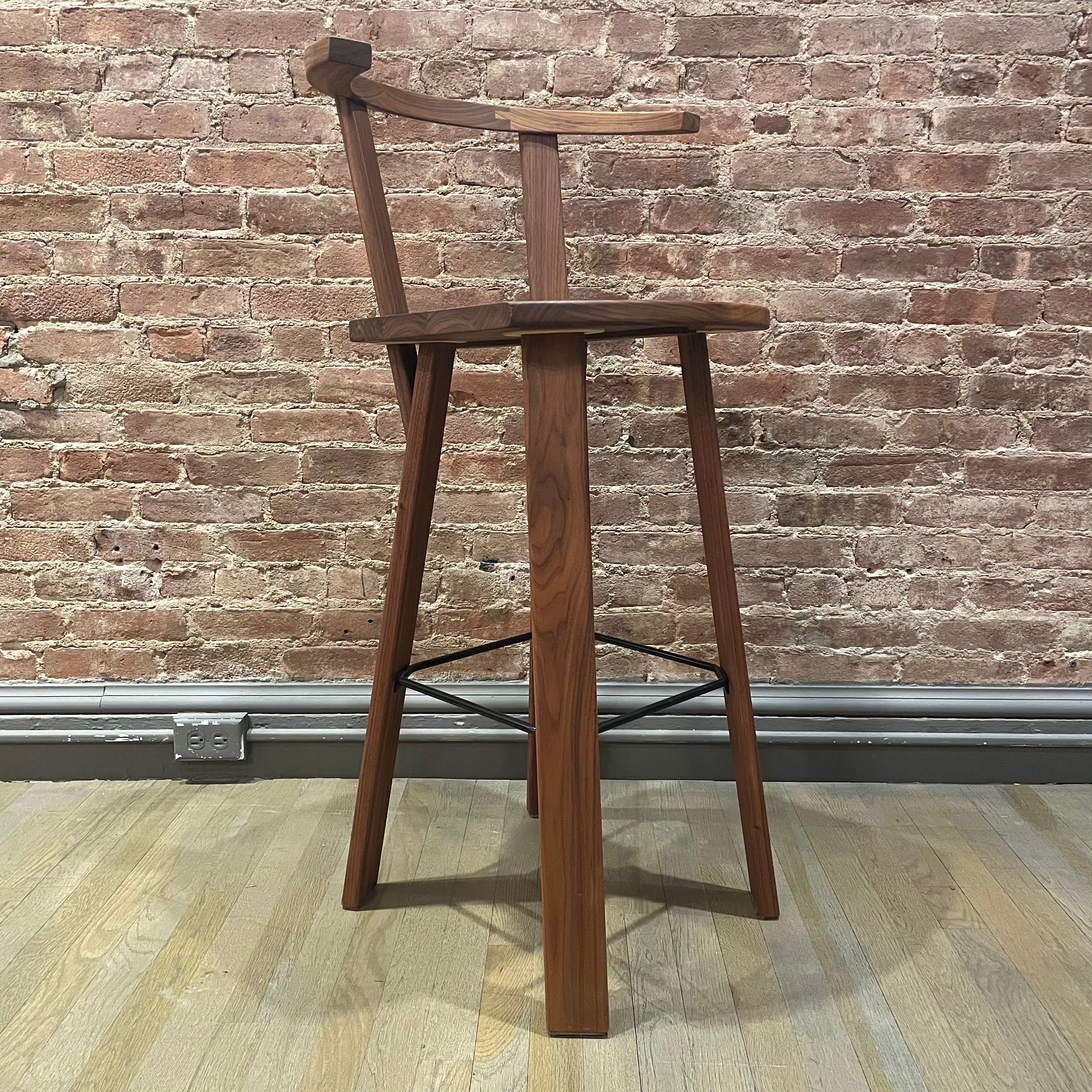 The Alpin Barstool with Armrest - Sale by Hussl embodies minimalist EOOS designs, standing on a wooden floor against an exposed brick wall. It features crossed metal support bars for style and stability in any space.