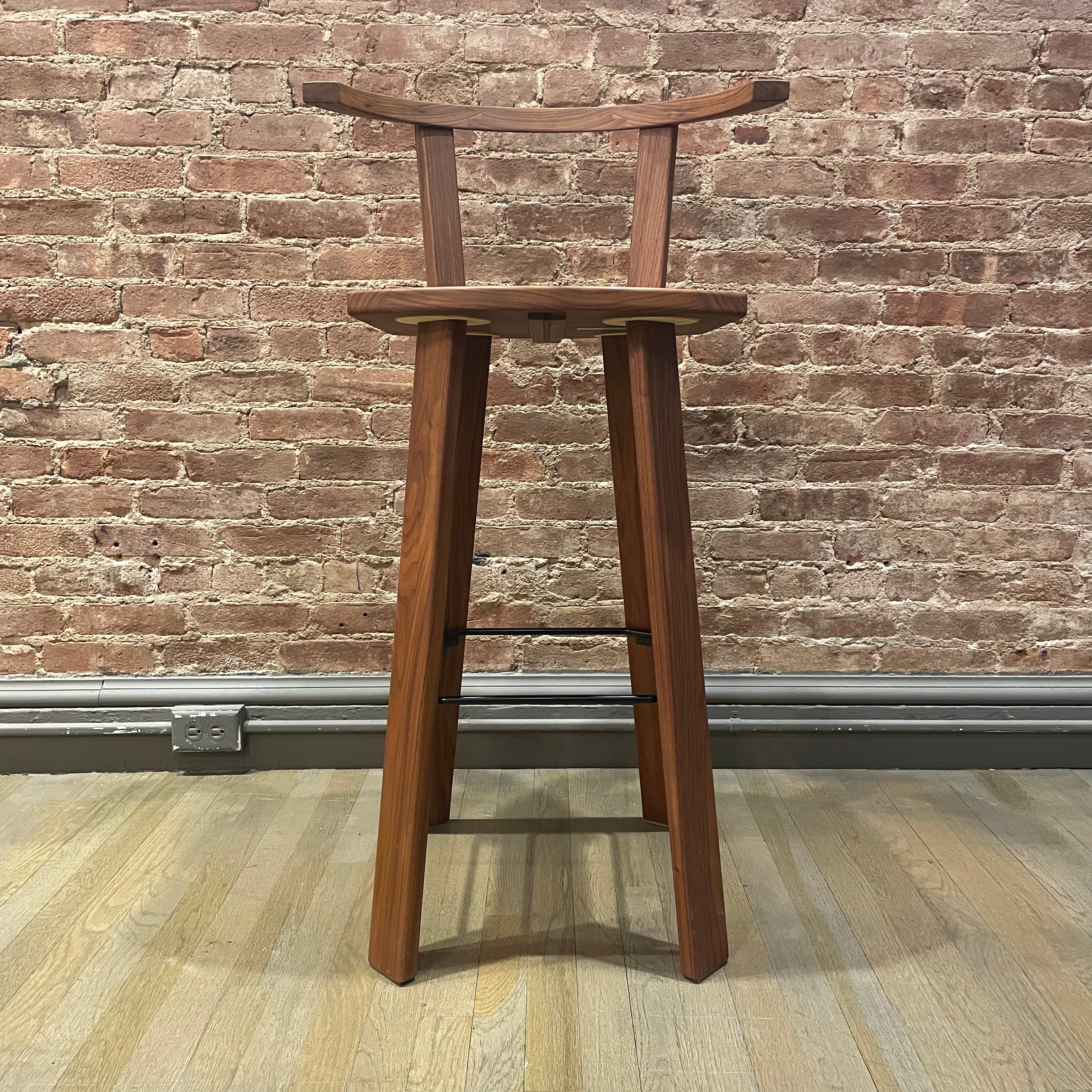 The Alpin Barstool with Armrest by Hussl, inspired by EOOS design, features a minimalist, high-back style with straight legs and a natural finish. Against a textured brick wall and set on a wooden floor, it resembles an Alpine bench.