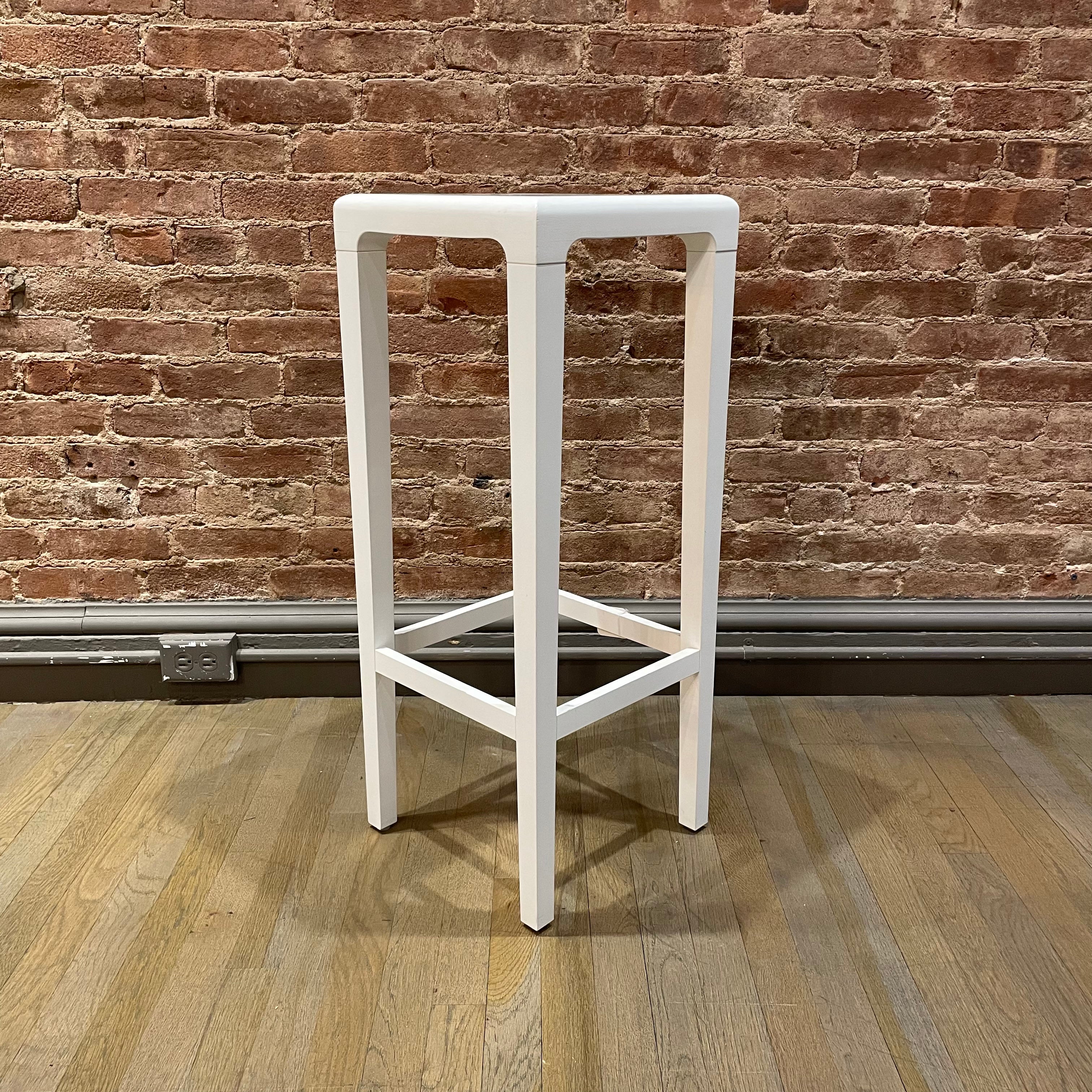 The Barstool Rioja - Sale by TON stands tall on a wooden floor, its white square design contrasting with a textured red brick wall. A gray baseboard completes this urban-chic look, available for NYC pickup.