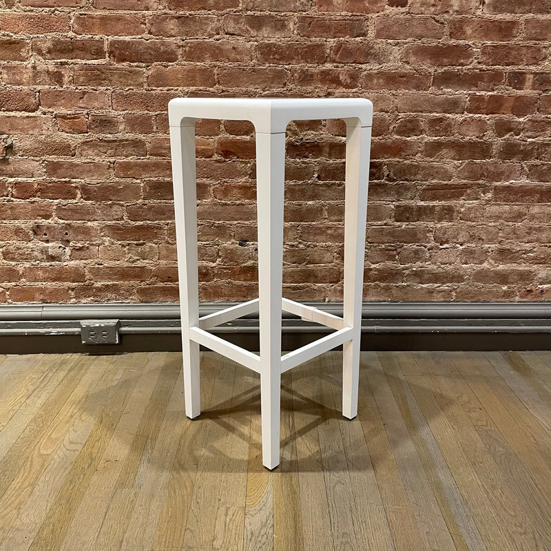 The Barstool Rioja - Sale by TON stands tall on a wooden floor, its white square design contrasting with a textured red brick wall. A gray baseboard completes this urban-chic look, available for NYC pickup.