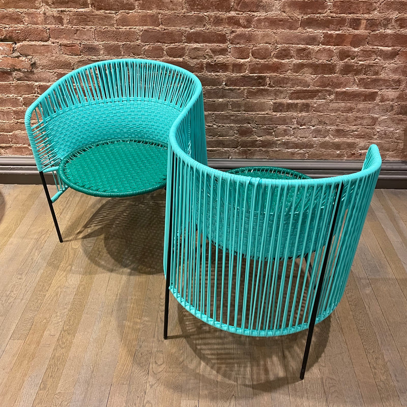 Two Caribe Double Chairs from Ames are positioned on a light wooden floor, featuring turquoise wicker with Momposino weave. They have thin black metal legs and sit against a textured brown brick wall.