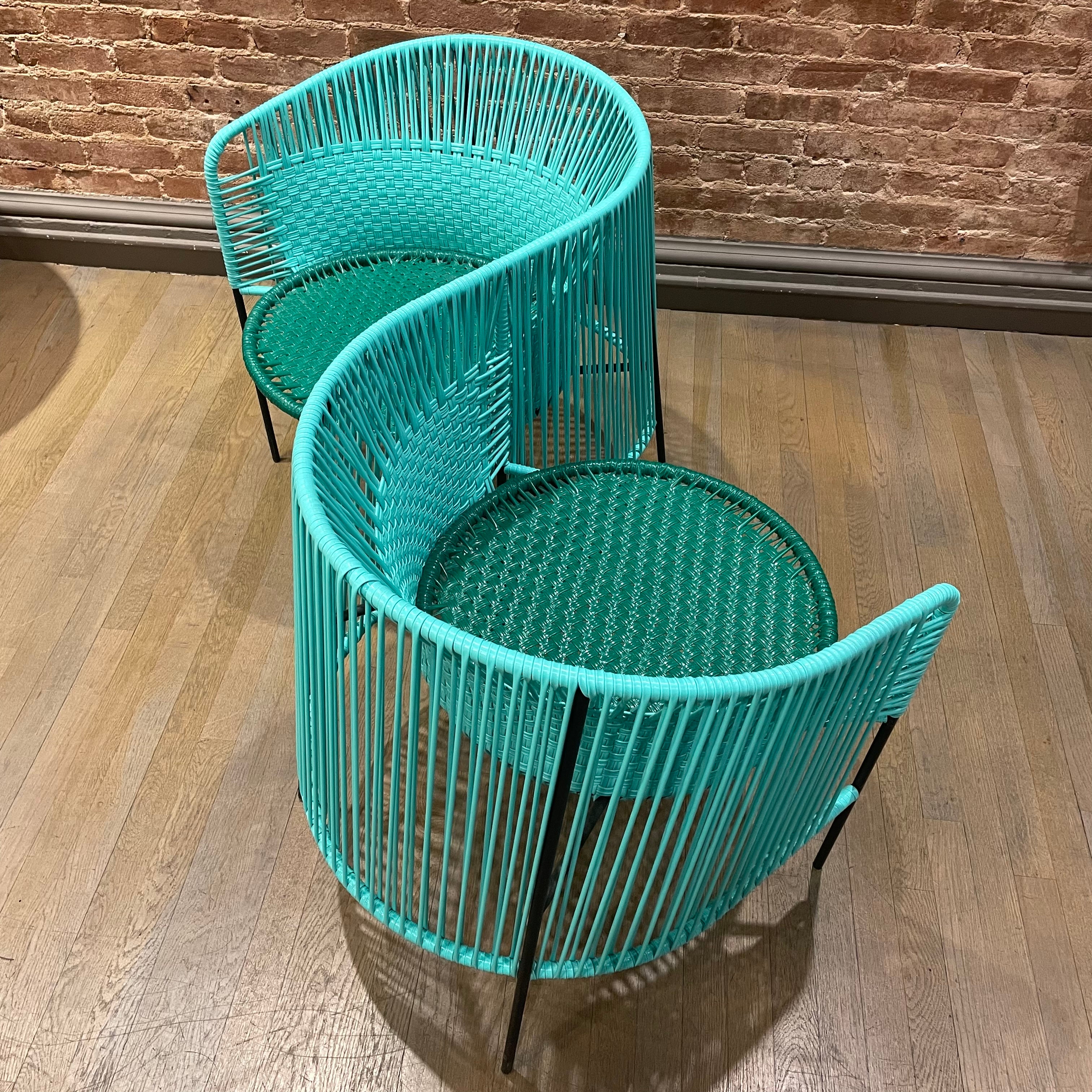 Two turquoise woven chairs from the Caribe collection by Ames, designed by Sebastian Herkner, feature circular seats in a mirrored S shape on a wooden floor with a textured brick wall backdrop. Crafted using the Momposino weaving technique. Available as the Caribe Double Chair - Sale.