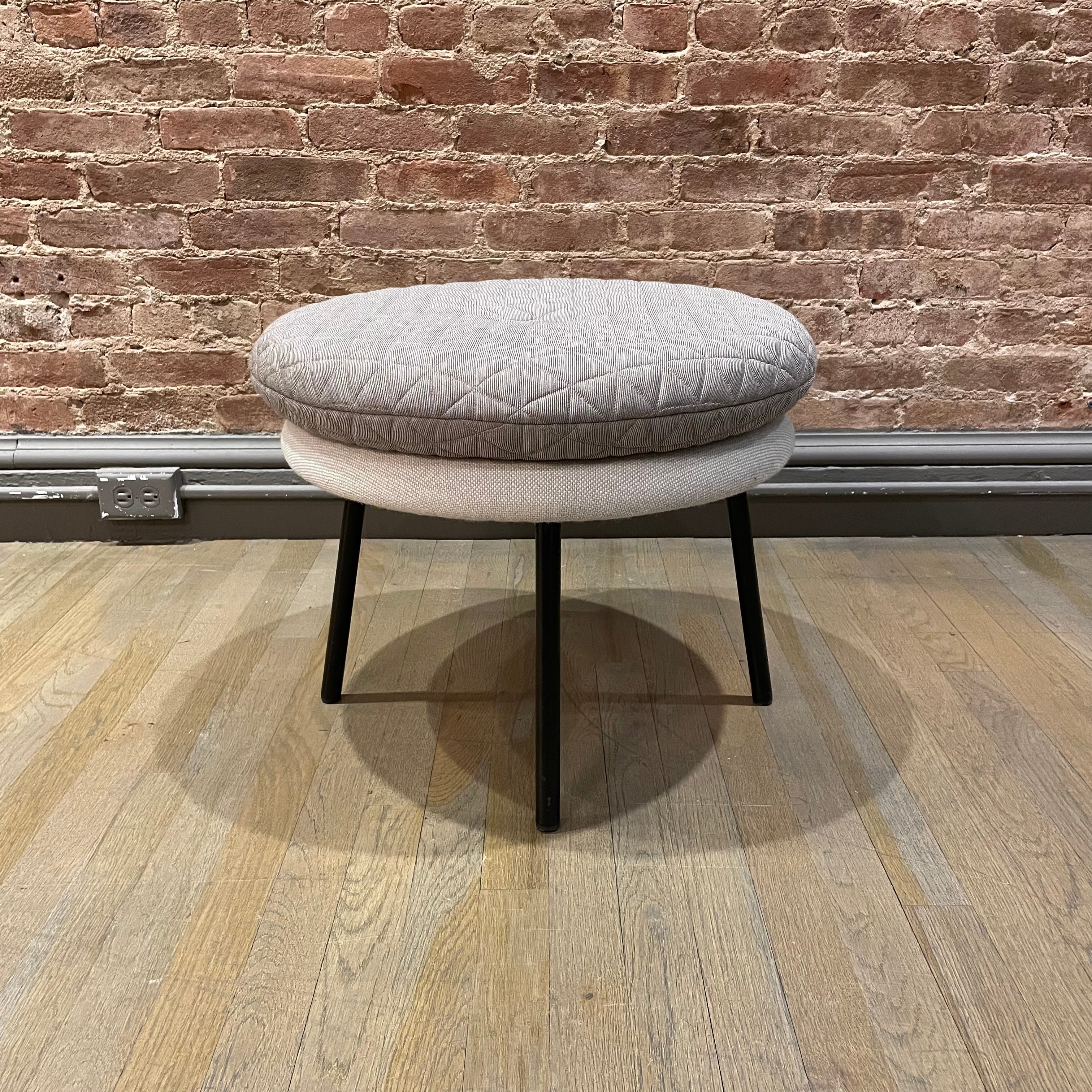 The Little Tom Stool by Richard Lampert, designed by Alexander Seifried, features a quilted gray upholstery and rests on thin black legs on a light wood floor. The setting includes a textured brick wall with an outlet at the base.