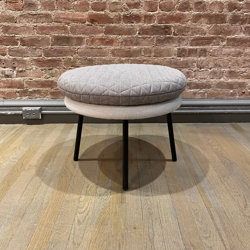 The Little Tom Stool by Richard Lampert, designed by Alexander Seifried, features a quilted gray upholstery and rests on thin black legs on a light wood floor. The setting includes a textured brick wall with an outlet at the base.