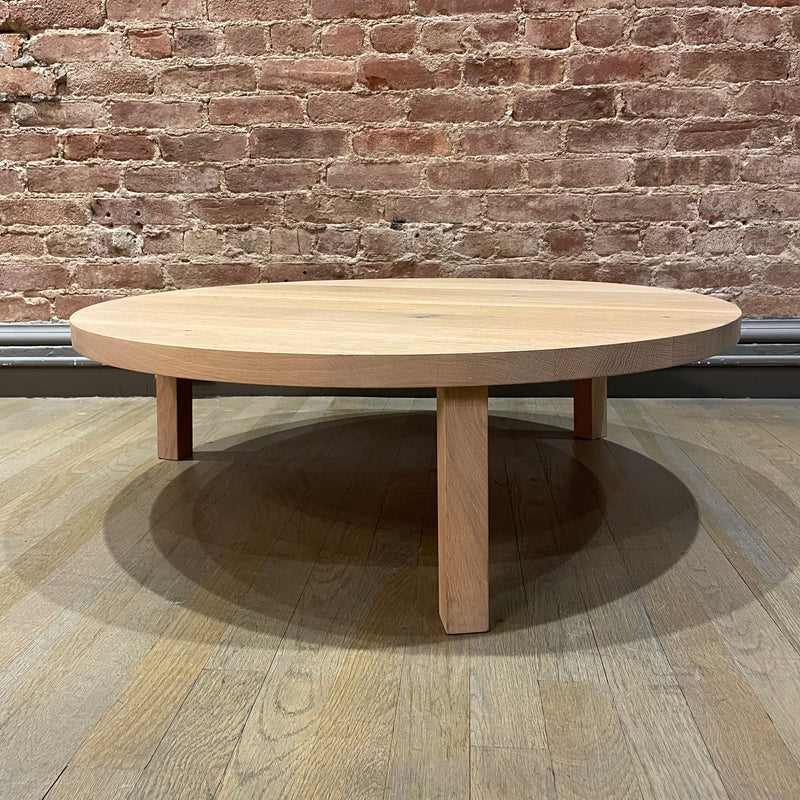 The PA05 LEILA coffee table by E15, crafted from solid European oak, graces the wooden floor. With its round shape, minimalistic design, and three legs, it stands elegantly against a brick wall backdrop, exuding modern simplicity.
