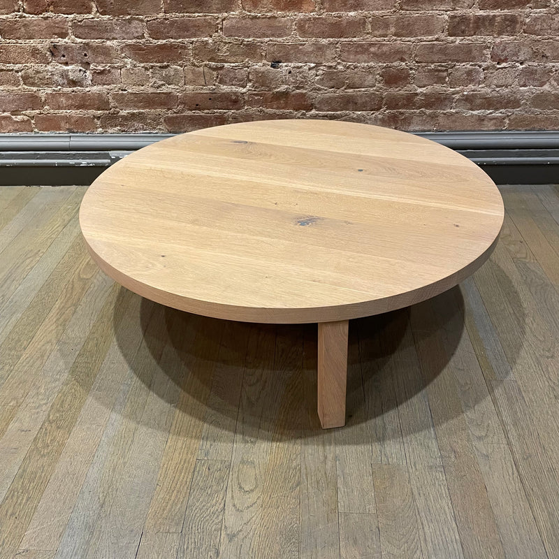 The PA05 LEILA by E15, a minimalist coffee table in solid European oak on wooden floors, boasts a light surface and three angled legs, enhanced by a brick wall backdrop.
