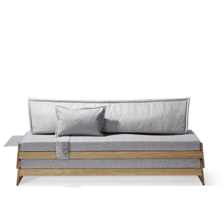 The Lönneberga Stacking Bed by Richard Lampert is a space-saving modern piece with a minimalist wooden frame and light gray cushions, including a back cushion and a matching throw pillow against a white background.