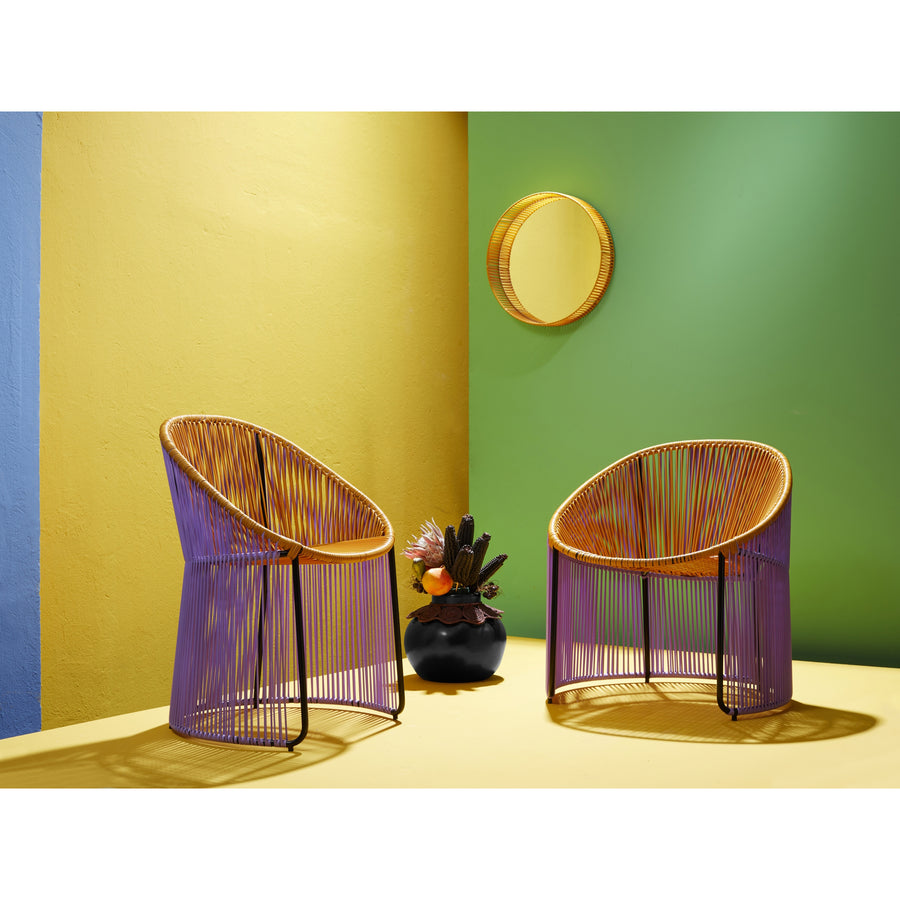 Two Cartagenas Lounge Chairs by Ames, featuring orange and purple woven designs, sit in front of a yellow wall. Adjacent is a green wall with a round wicker mirror. On the vibrant yellow floor, a black vase filled with exotic flowers completes the colorful scene.