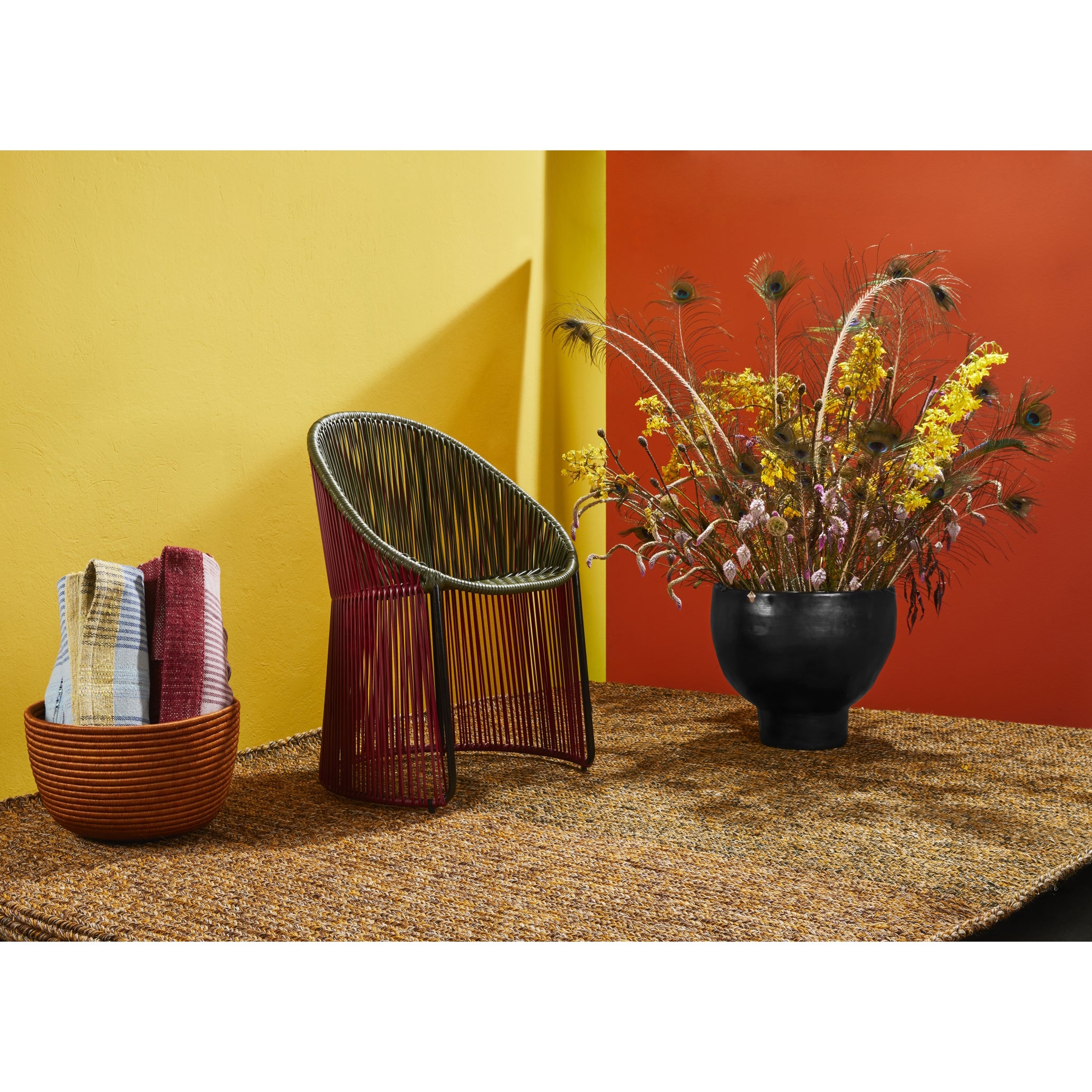 A modern room scene features the Ames Cartagenas Dining Chair, a large black vase with dried flowers and peacock feathers, and a woven basket of towels on a textured rug set against bright yellow and orange walls.
