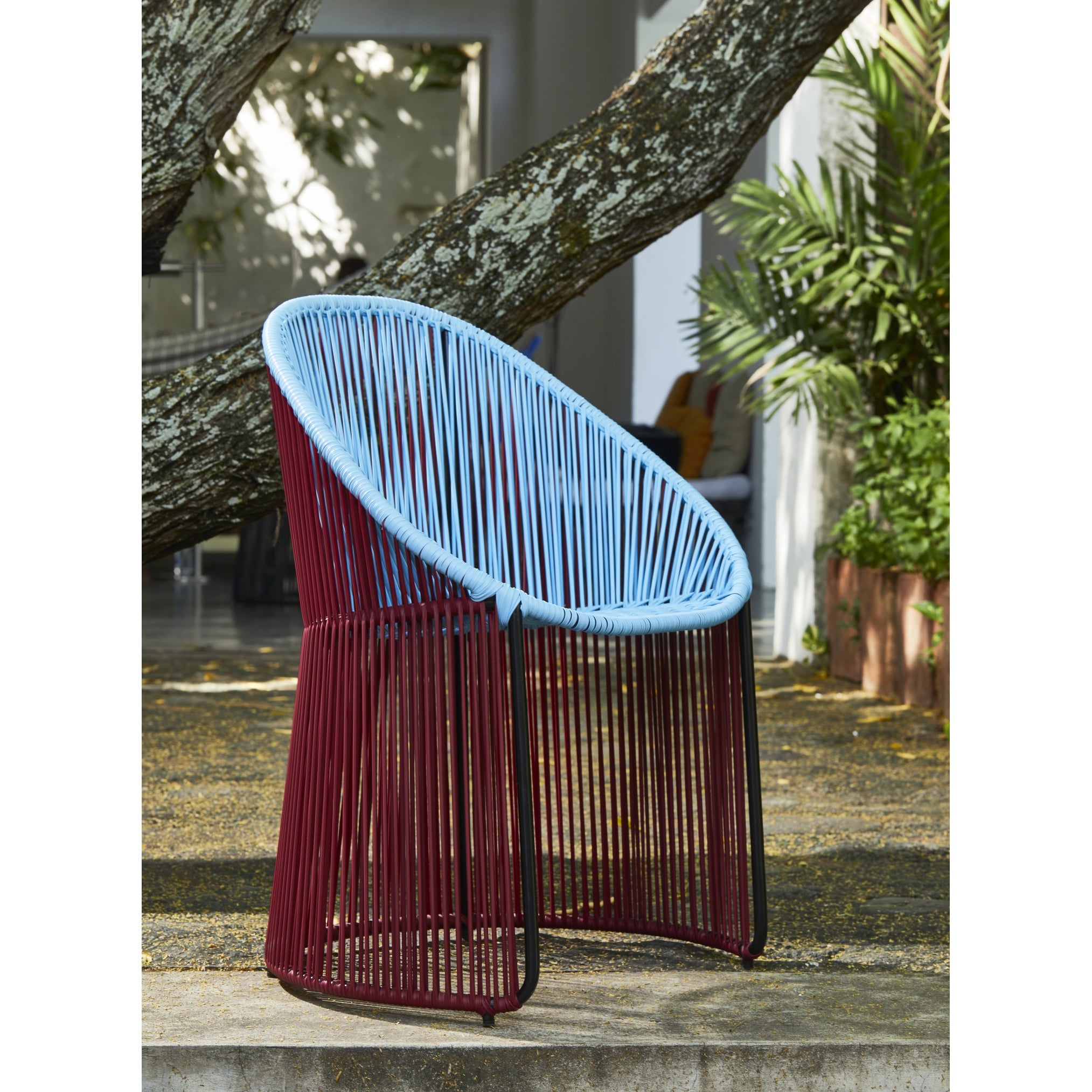The Ames Cartagenas Dining Chair by Ames, featuring maroon legs and a sky blue woven backrest, is placed on a concrete patio beneath a large tree with greenery and a building in the background.