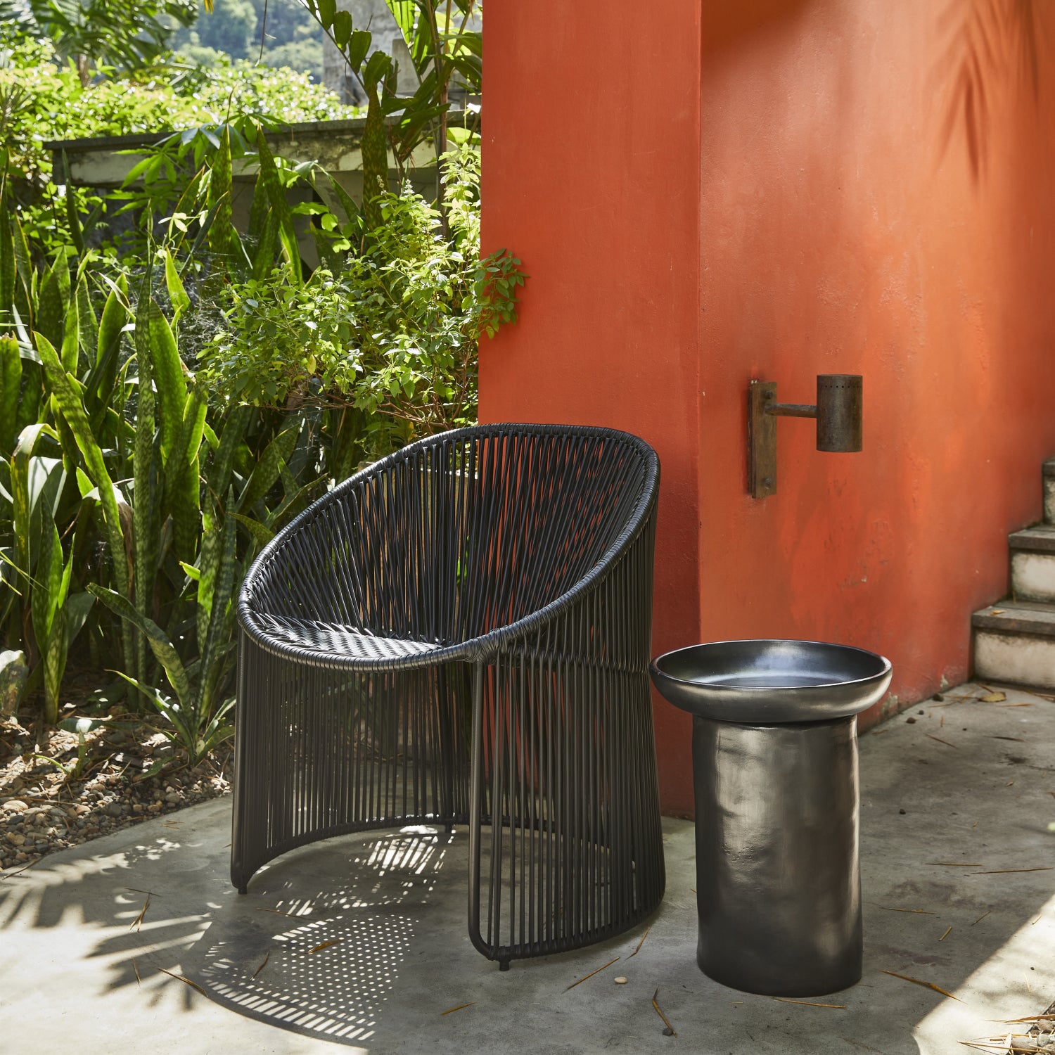 A modern outdoor scene showcases a chic black Cartagenas Lounge Chair by Ames, paired with a matching round black side table. This setup is placed on a concrete patio, framed by a vibrant orange wall and lush green plants.