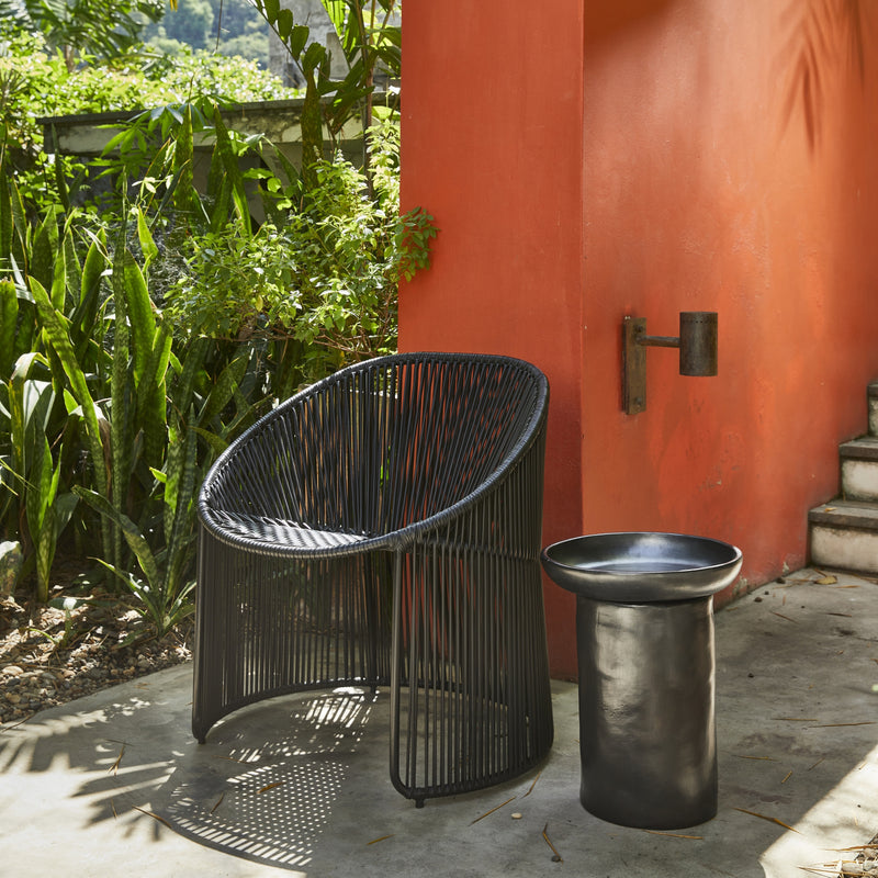 A modern outdoor scene showcases a chic black Cartagenas Lounge Chair by Ames, paired with a matching round black side table. This setup is placed on a concrete patio, framed by a vibrant orange wall and lush green plants.