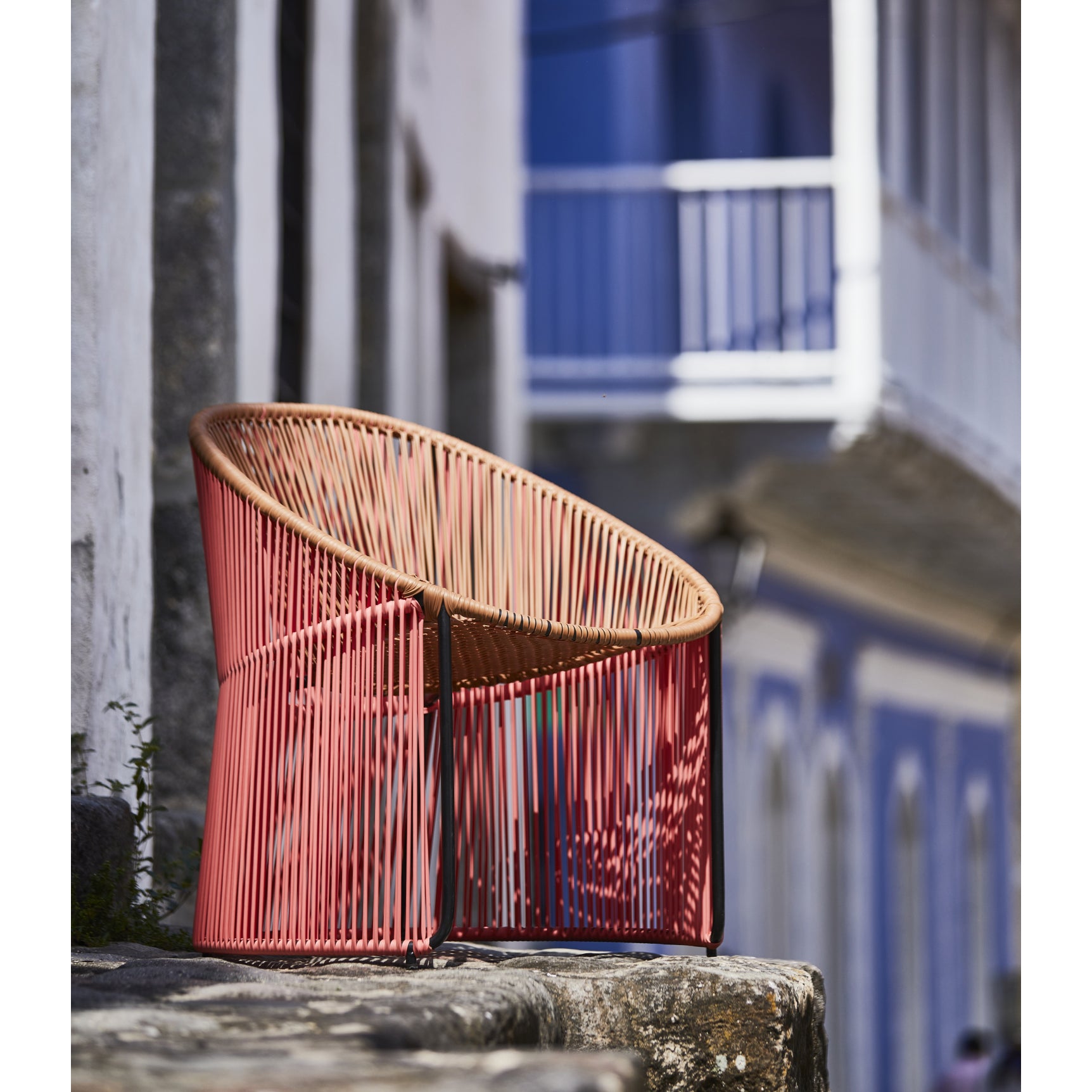 The Cartagenas Lounge Chair from Ames brings modern flair with its striking red wire frame and brown seat, set on a stone terrace. A white and blue building with a balcony subtly blurs in the background, enhancing the serene outdoor setting.
