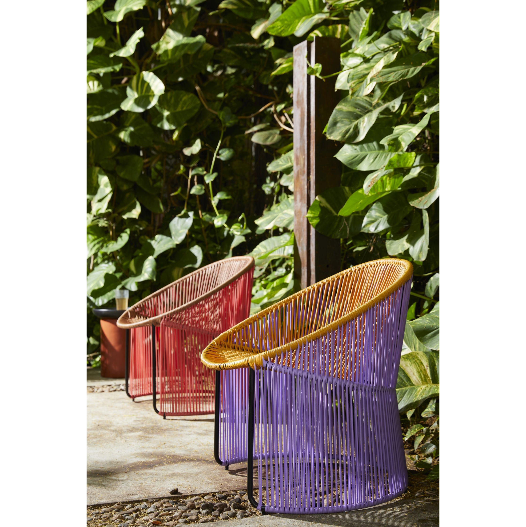 Two Cartagenas Lounge Chairs by Ames—one purple with a yellow seat and the other red—are placed on a stone pathway, surrounded by lush green foliage, creating a vibrant and inviting outdoor setting.