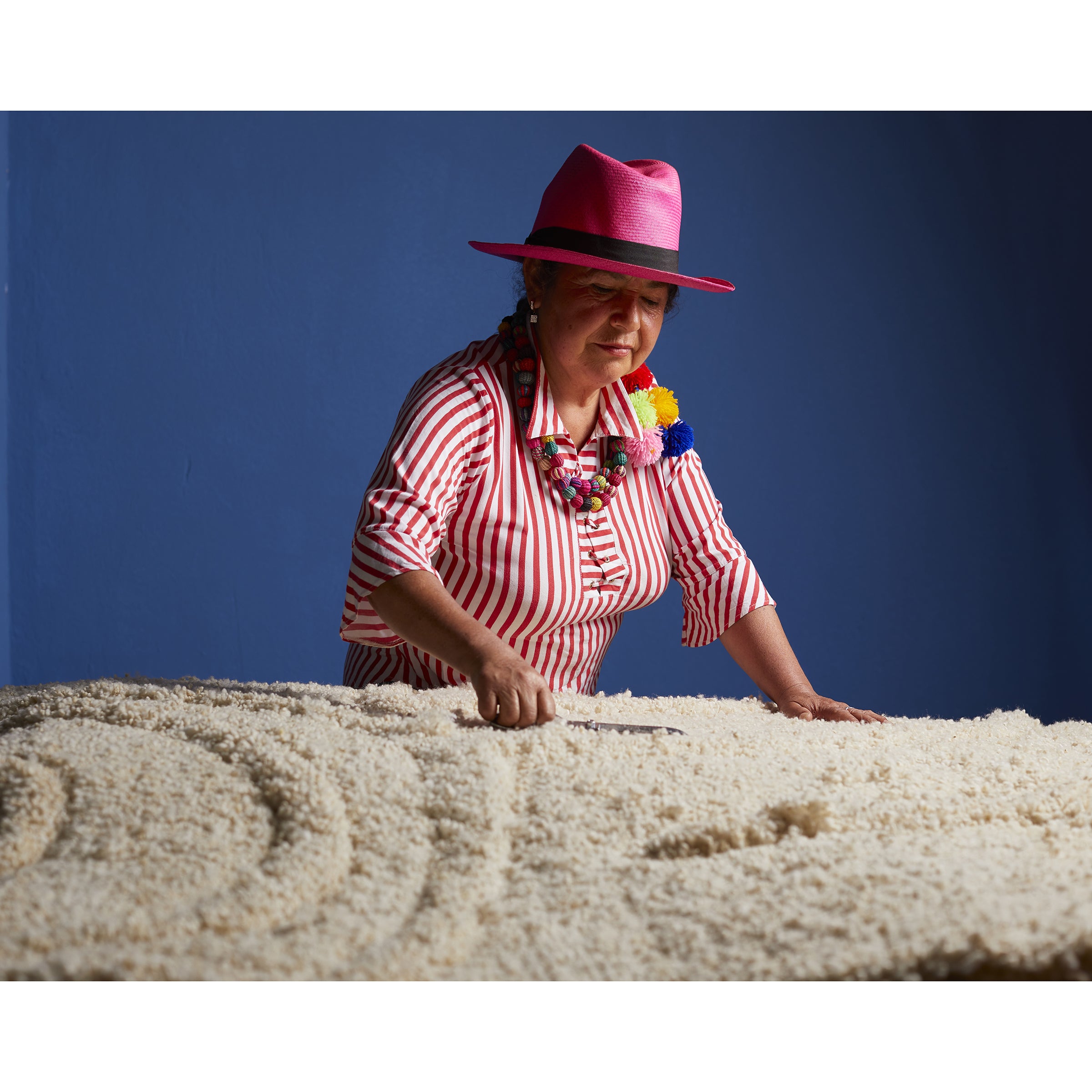 Against a blue backdrop, an individual in a pink hat and striped shirt arranges grains in patterns that evoke the Zenues Rug by Ames.