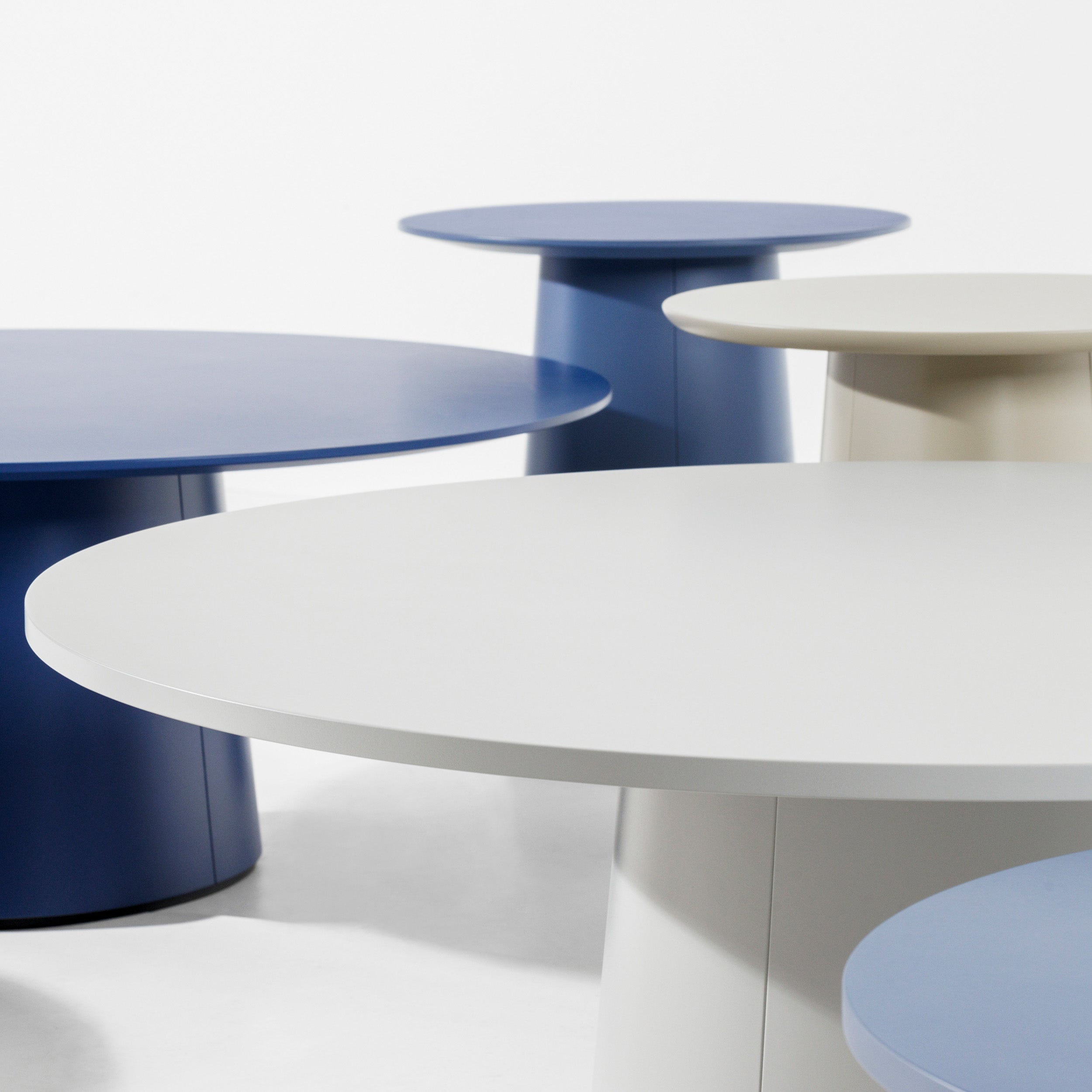 The P.O.V. Coffee Table 473 by TON is a modern round table with a cylindrical base and a smooth, minimalist tabletop. It features matte finishes in blue, white, or gray and is displayed on a white background in various sizes.