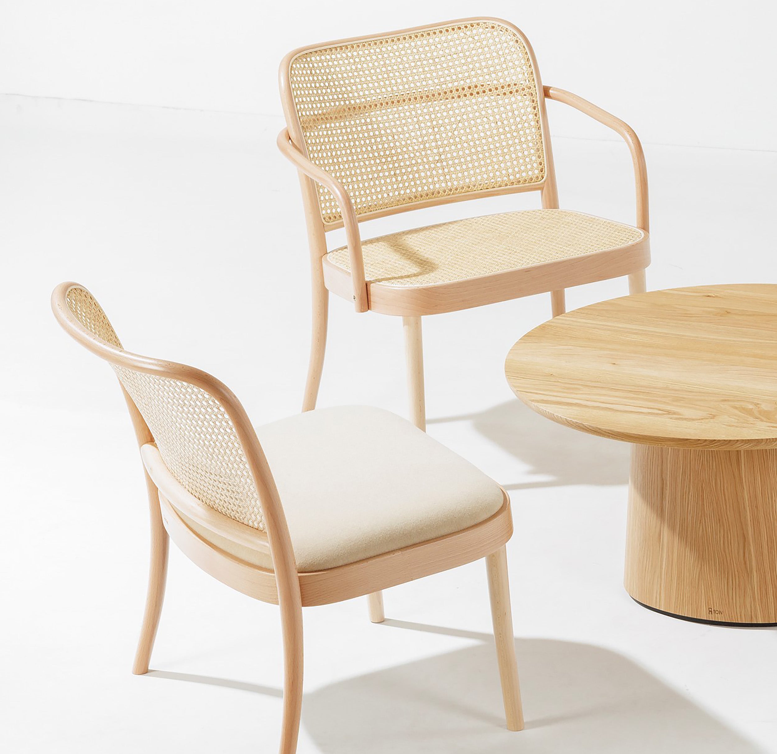The Lounge Armchair 811 by TON, featuring light wood with cane weaves and neutral cushions, is arranged around a round wooden table on a white floor. The design showcases smooth curves and a natural finish, embodying the chic lounge armchair style.