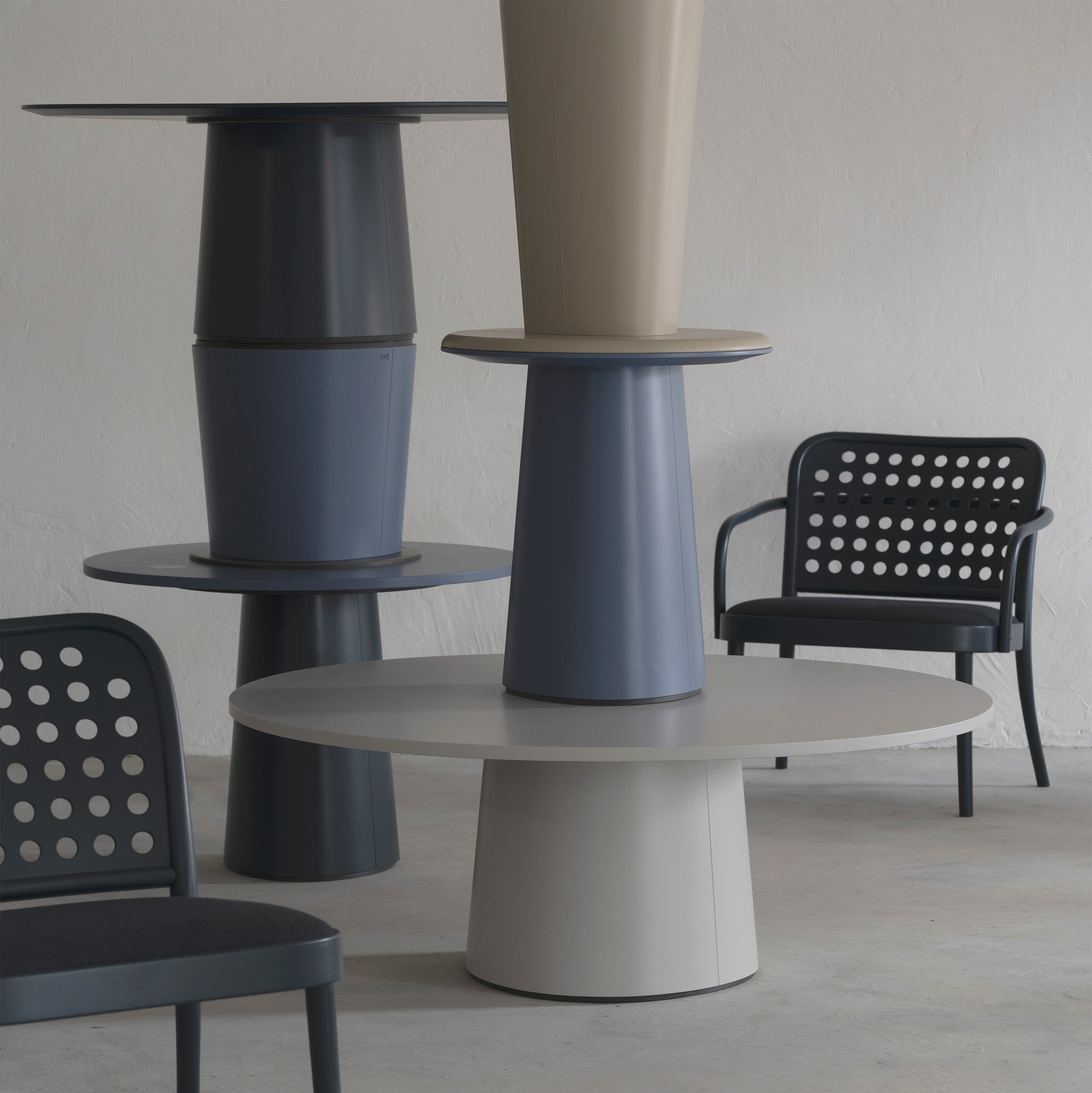 The modern interior features stacked, column-like P.O.V. Coffee Tables 474/475 by TON in dark green, blue, and beige. Two dark green chairs with perforated backs complement the tables against a light gray wall and floor for a minimalist yet stylish appeal.