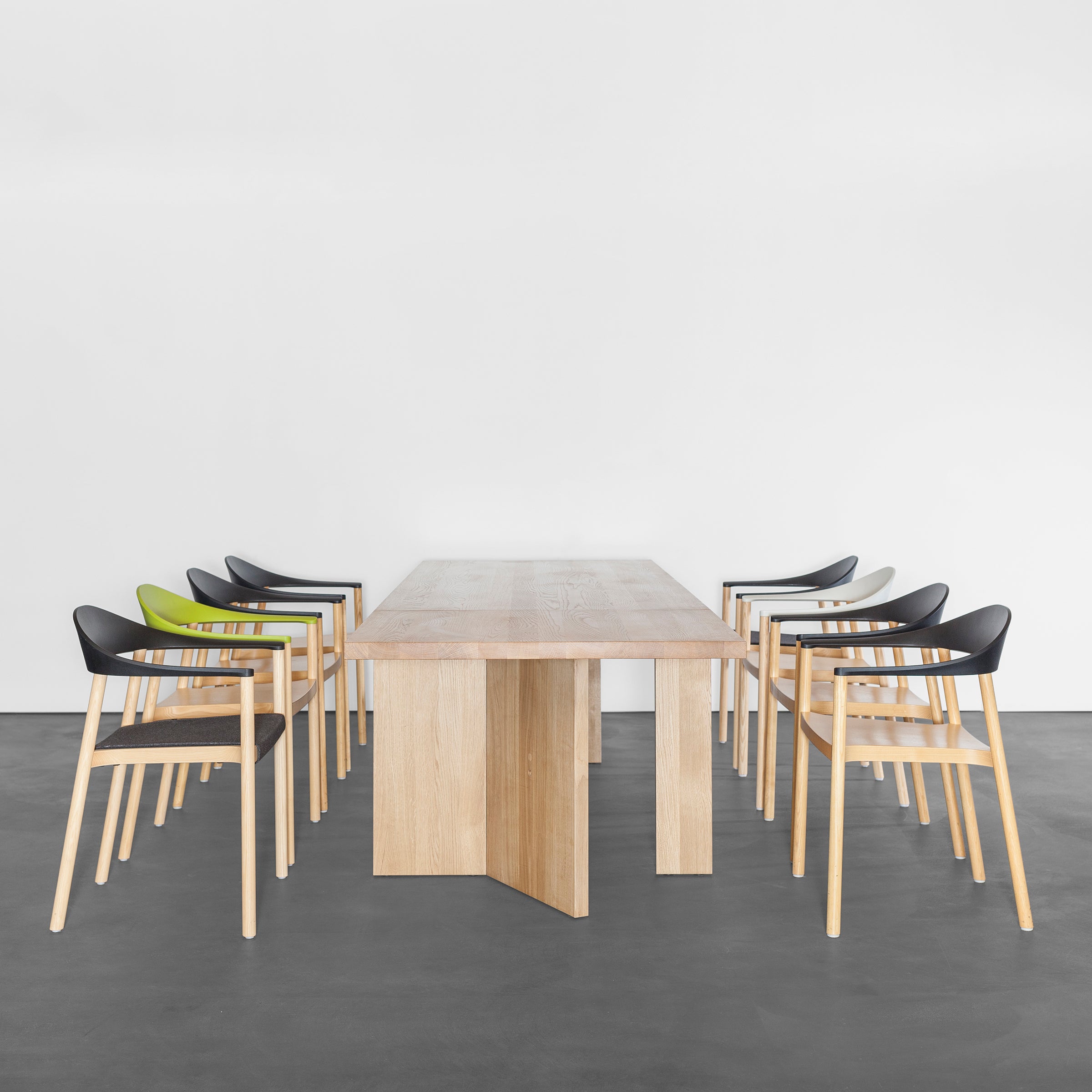 A minimalist dining room showcases a Sanktjohanser Alphabet Folding Table with solid wood finishes and ten modern chairs. Two chairs have lime green seats, while the others are black. The dark floor and plain white backdrop enhance the clean, open feel.