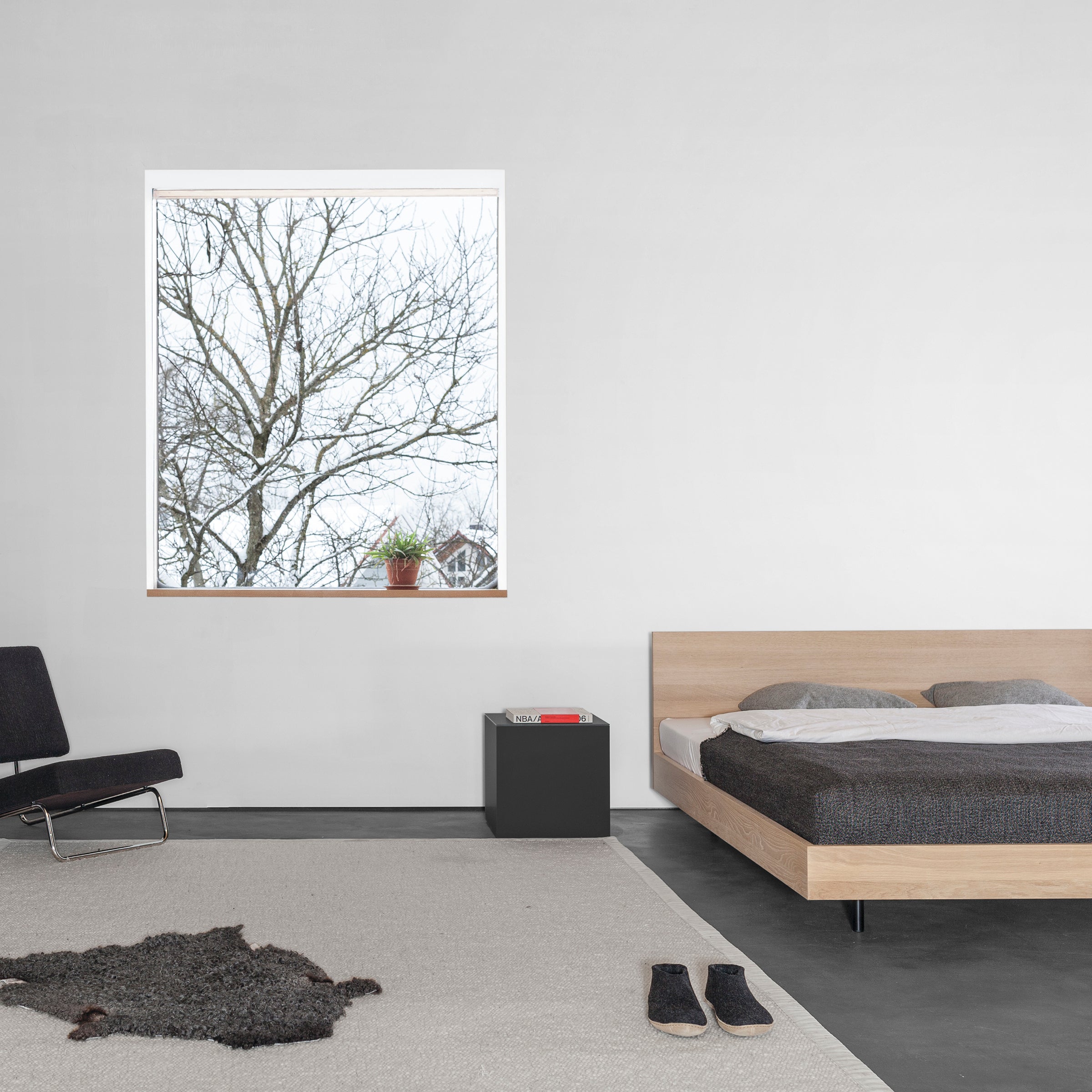 The minimalist bedroom features the Iku Bed Series by Sanktjohanser placed against a window with bare trees. A small side table holds a book, next to a black chair and gray rug. Nearby slippers and a potted plant add life to the neutral palette.