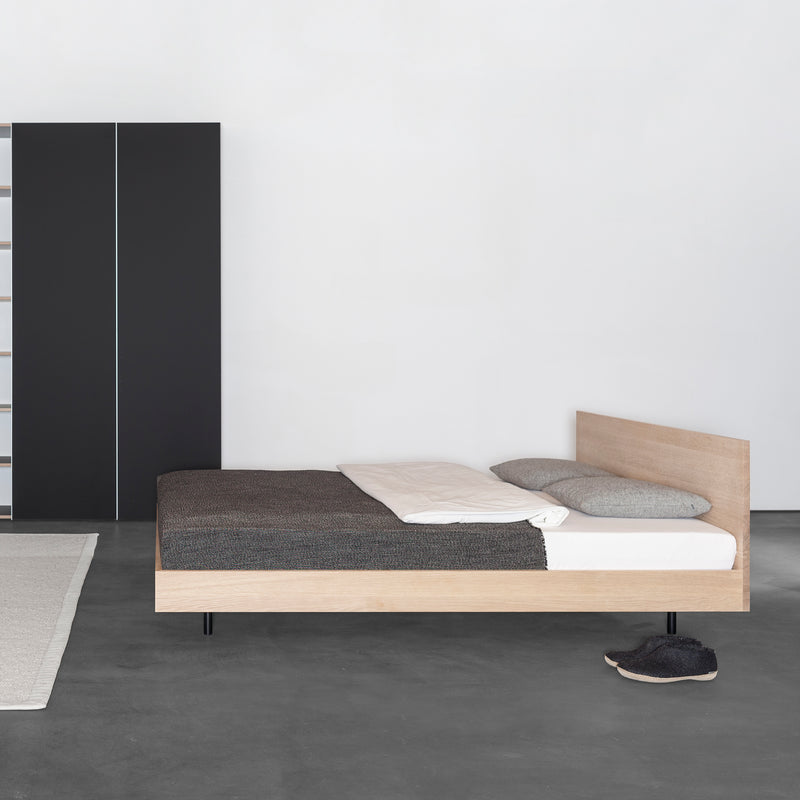 A minimalist bedroom showcases the Sanktjohanser Iku Bed Series with light solid wood and gray bedding. A single pillow adorns the bed, gray slippers lie on the concrete floor, a dark wardrobe contrasts against a white wall, and part of a light rug peeks out beneath the stackable beds base.