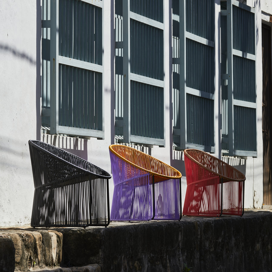 Three vibrant Cartagenas Lounge Chairs—black, purple, and red—from Ames are arranged in front of a white wall with blue windows. Perched on a stone ledge, these chairs cast shadows under the bright sunlight.