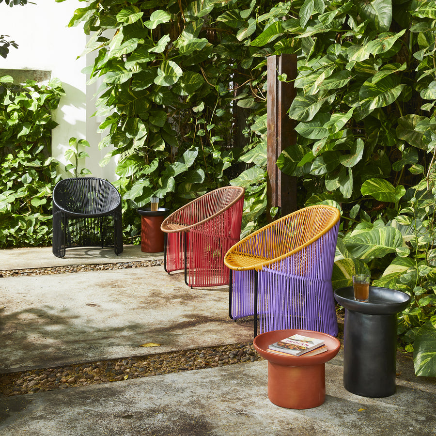 Colorful Ames Cartagenas seating, including three dining chairs in yellow, pink, and purple, plus a black Cartagenas Lounge Chair, are arranged amidst lush green plants. Two cylindrical side tables hold drinks and books, enhancing the cozy garden setting.