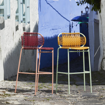 Two vibrant Ames Cielo Barstools by Ames, one in red and one in yellow, stand on a cobblestone street with white walls and a blue-painted background, creating a bright summer outdoor scene.
