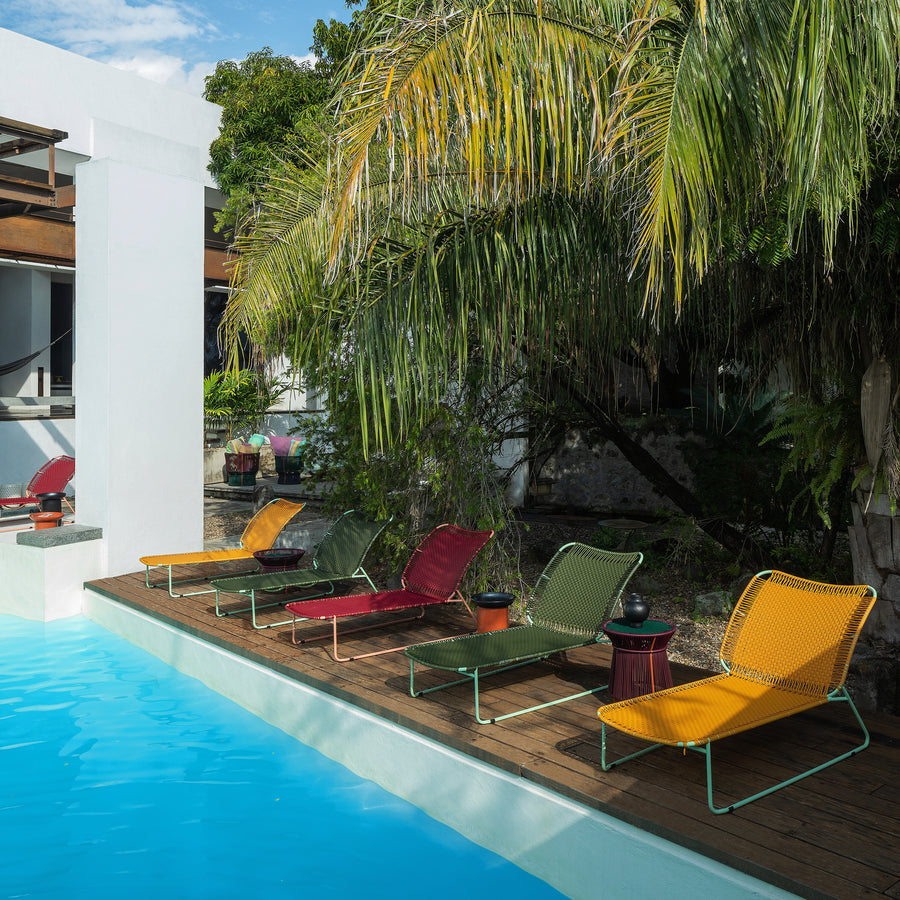 A row of colorful lounge chairs and the Ames Cielo Daybed by Ames rest on a wooden deck beside a bright blue pool, surrounded by lush palm trees and modern white buildings.