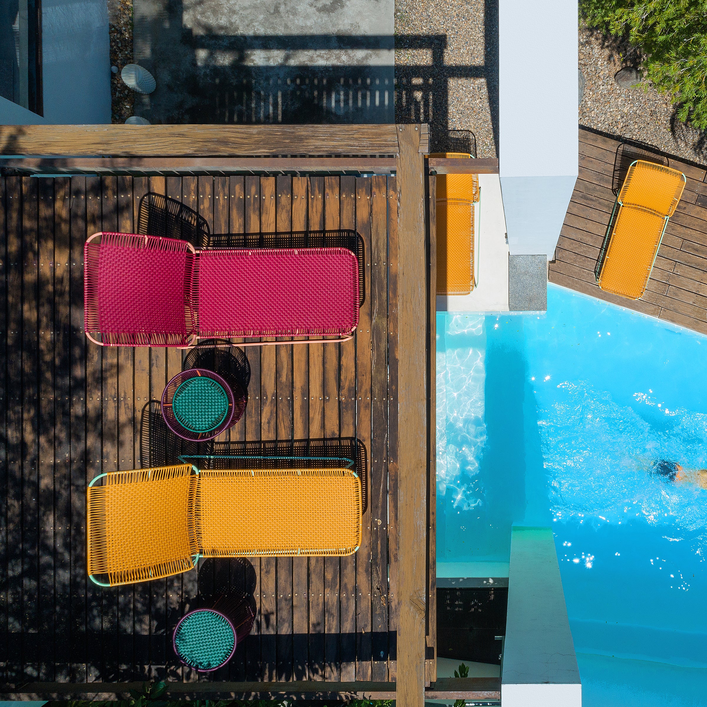 Aerial view of a wooden deck with colorful woven lounge chairs, the Ames Cielo Daybed by Ames, and small tables beside a bright blue pool where someone is swimming, framed by shadows and lush greenery.