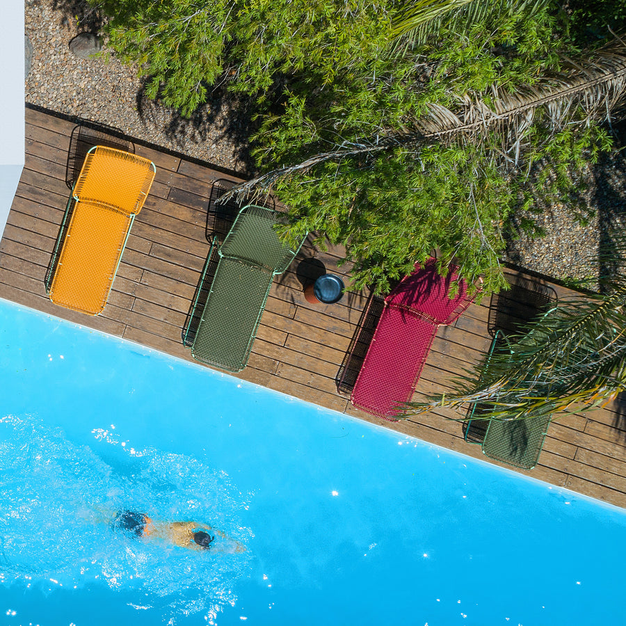 A person swims in a bright blue pool next to a wooden deck with colorful lounge chairs, lush greenery, and an Ames Cielo Daybed by Ames, seen from above.