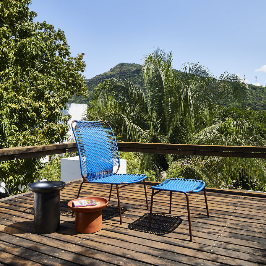 A blue chair with the Ames Cielo Ottoman by Ames, inspired by Sebastian Herkner, sits on a wooden deck with side tables and a magazine, surrounded by lush green trees and distant mountains under a clear blue sky.