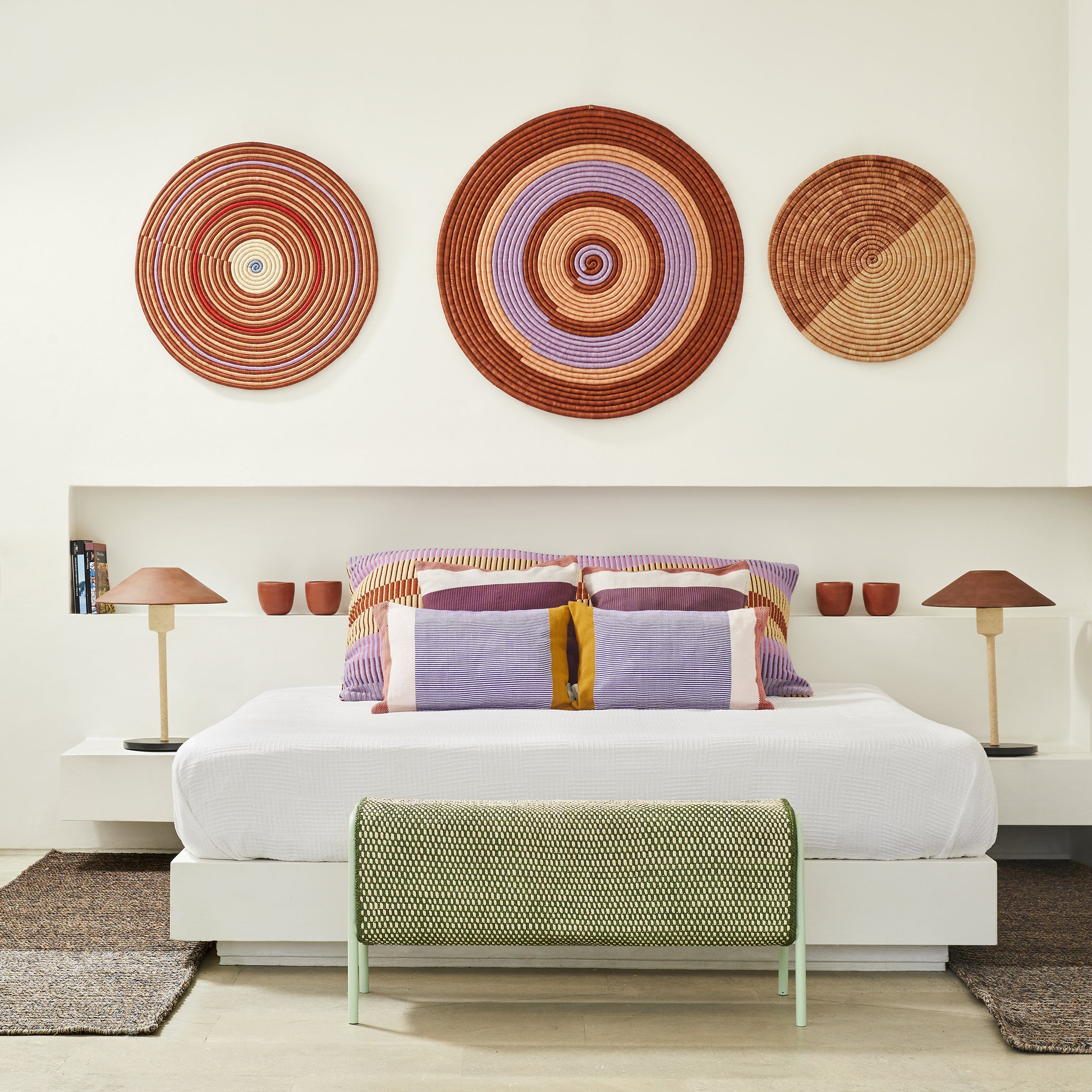 A modern bedroom showcases a white bed adorned with colorful pillows. Above, three circular woven wall decorations highlight Colombian craftsmanship in red, orange, and purple. Matching Ames Raiz Table Lamps with ceramic shades and a green bench complete the setup.