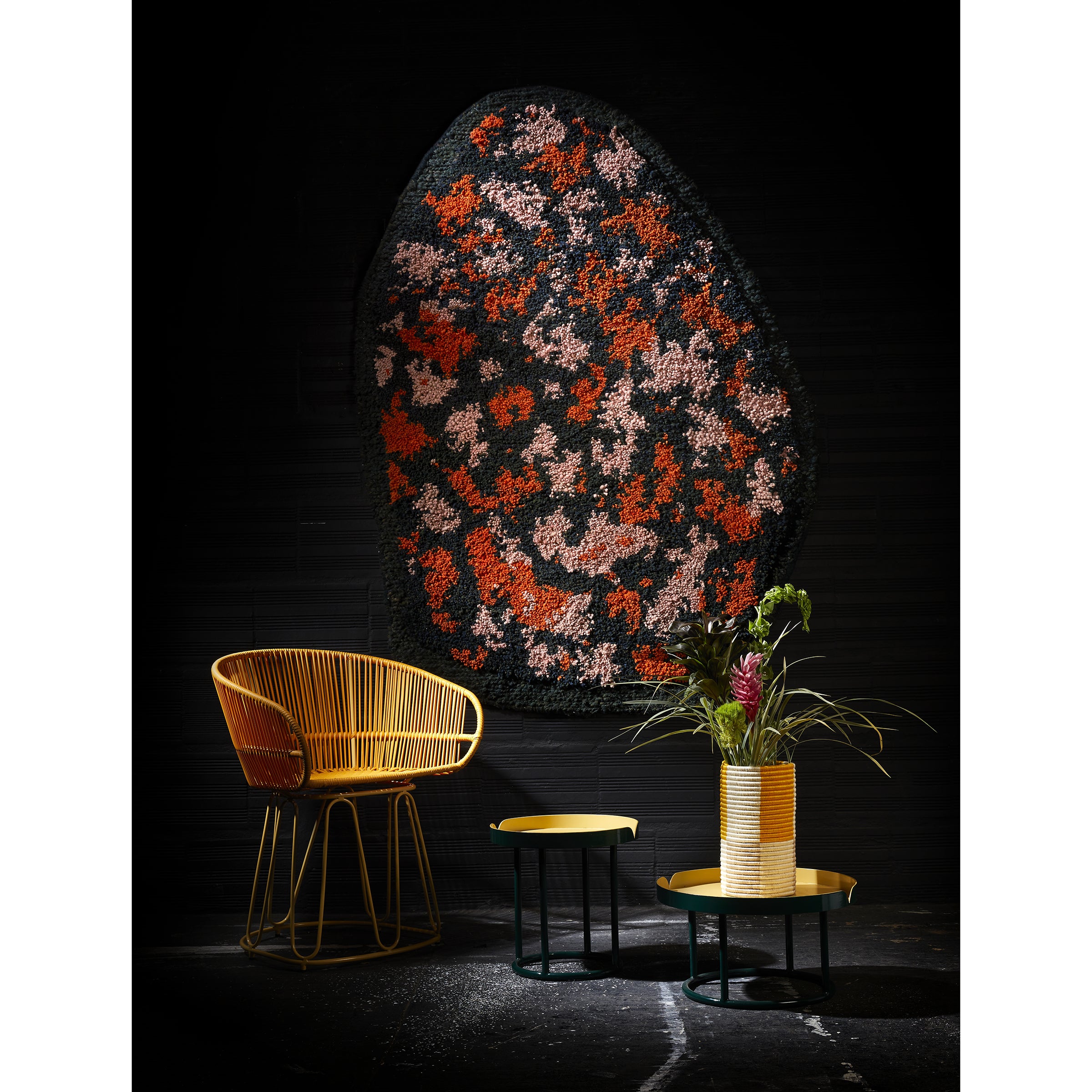 A modern interior features a yellow woven chair, two round tables (green and gold), a potted plant, and a bold Ames Zipa Rug by Ames, designed by Sebastian Herkner, hanging as colorful wall art.