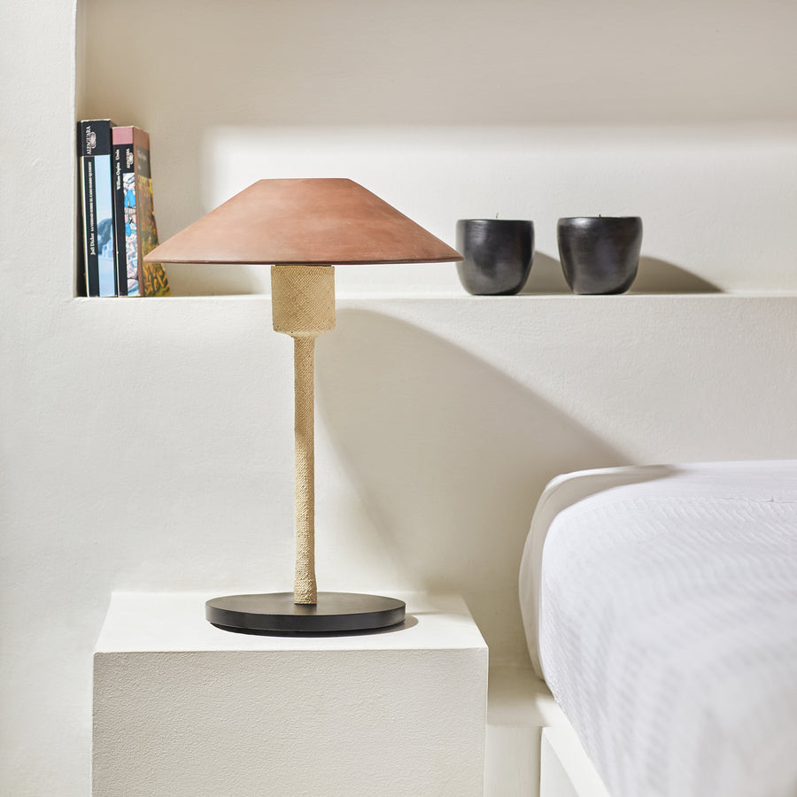 The Raiz Table Lamp by Ames, featuring a ceramic shade, adorns a white bedside table. In the background on a shelf against the white wall are two black cups and some books. Part of the bed with a white duvet, showcasing exquisite Colombian craftsmanship, is also visible.